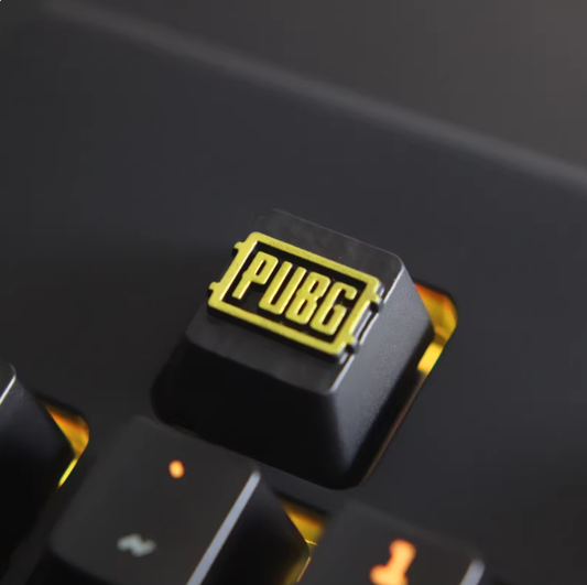 PUBG logo metal keycap, black base, gold embossed logo, installed on mechanical keyboard, warm yellow backlight, close-up.