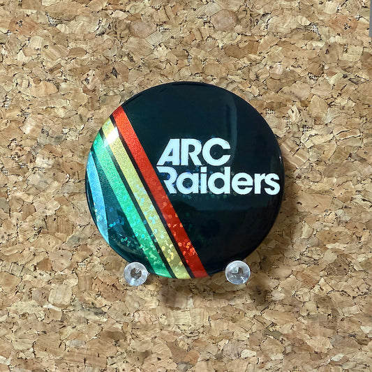 Round ARC Raiders pin button featuring the white text logo with retro green, yellow, and red stripes on a black background, finished with a holographic glitter effect.