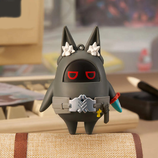 This is a dark gray figurine with pointed ears adorned with white star-shaped accents. It features a black faceplate with red U-shaped eyes and a silver tactical belt across its chest. The figurine is placed on a beige woven-texture surface, with a blurred desk and keyboard in the background.