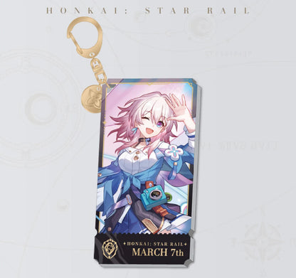 This Honkai: Star Rail acrylic keychain features March 7th in a cheerful, playful pose, with a golden clasp and chain, set against a soft pink background.
