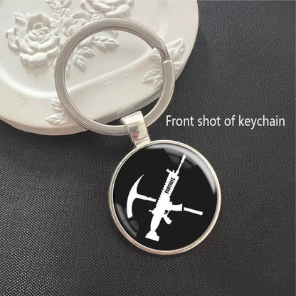 Fortnite-themed keychain with crossed pickaxe and rifle silhouette on a black background. Set in a polished silver frame with a scratch-resistant dome, ideal for keys or backpacks.