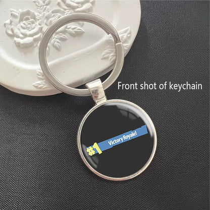 Fortnite-themed keychain featuring the iconic "#1 Victory Royale!" text on a black background. Set in a polished silver frame with a scratch-resistant dome, perfect for keys or backpacks.