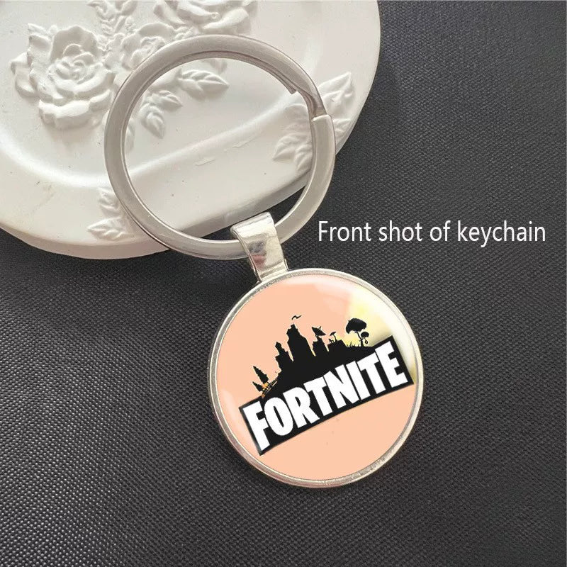 Fortnite keychain with the iconic game logo and skyline silhouette, set against a soft peach background. Encased in a polished silver frame with a scratch-resistant dome, perfect for keys or backpacks.