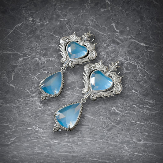 A pair of ornate silver earrings with sculpted heart-shaped studs and teardrop pendants, both set with shimmering light blue gemstones. whose unique texture creates a subtle contrast in the light.