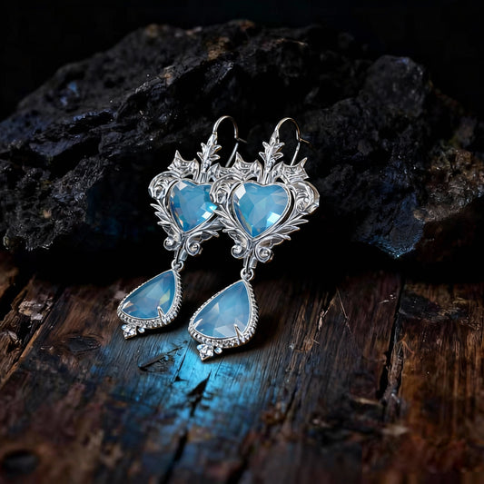 A pair of ornate silver earrings with sculpted heart-shaped studs and teardrop pendants, both set with shimmering light blue gemstones. whose unique texture creates a subtle contrast in the light.