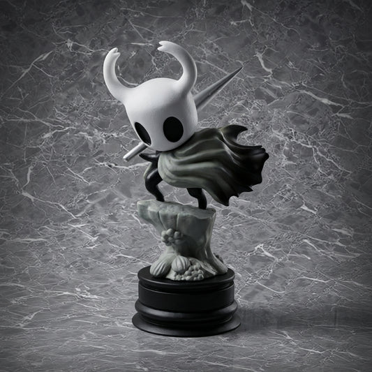 This is a detailed figurine of a character from the game "Hollow Knight," featuring a white masked head with large black eyes, a flowing gray cape, and standing on a rocky base, perfect for collectors and fans of the game.