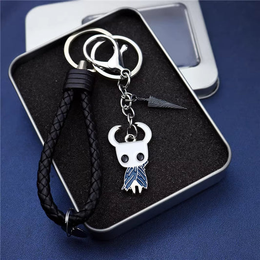 This is a sleek keychain featuring a braided black leather strap, a metal ring, and a detailed charm of a character with horns and a blue cape, along with a small knife - like accessory, all presented in a box.