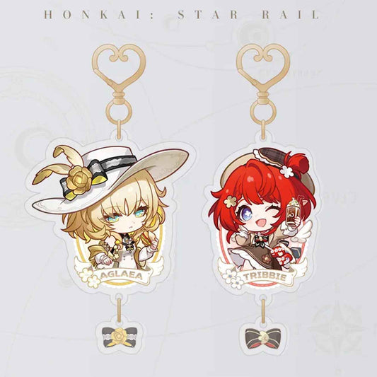 This image showcases two chibi-style acrylic keychains from Honkai: Star Rail. On the left is Aglaea, a blonde girl in a white hat with a gold rose, and on the right is Tribbie, a red-haired girl holding a drink. Both have heart-shaped clasps and small bow pendants.